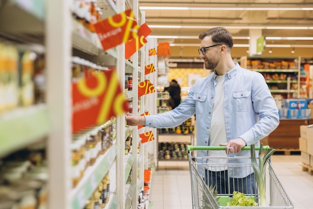 Man in supermarket aisle pushing trolley and looking at prices.